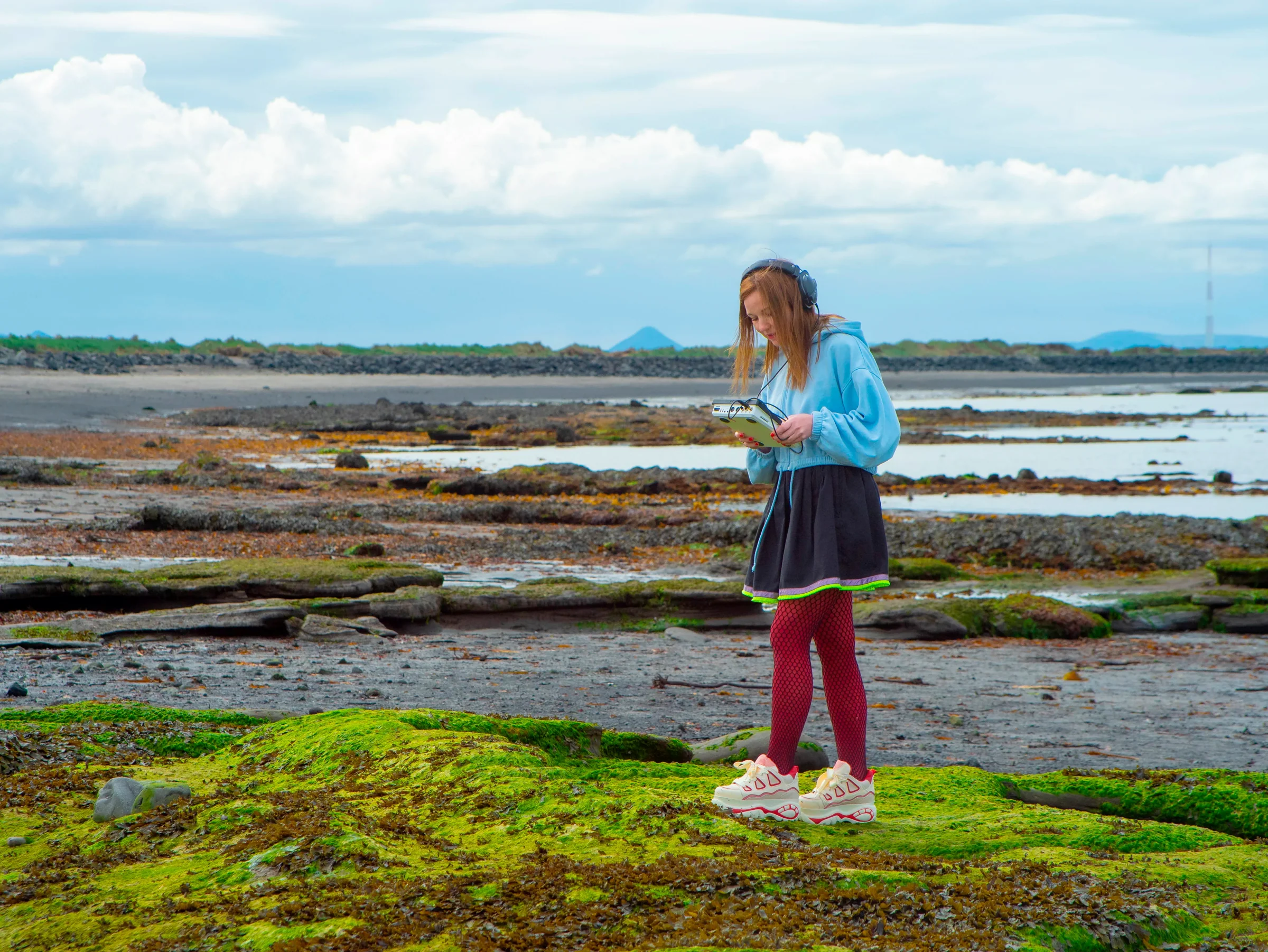 A musician playing FIRST LOVE with headphones on a moss-covered rocky shoreline.